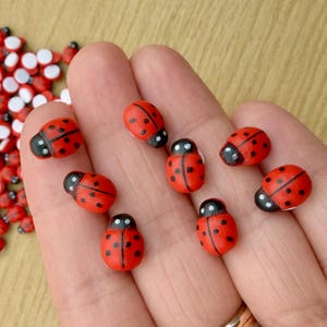 May include: A collection of small, rounded ladybird-shaped beads featuring a bright red body with black spots and a black head. The ladybirds are painted with a smooth finish and include a central line down the back, mimicking the appearance of wings.