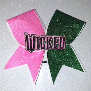 Peut inclure: Un nœud de pom-pom girl scintillant avec un design divisé. Un côté est rose vif, l'autre est vert foncé. Le mot "WICKED" est imprimé en noir avec un contour rose au centre du nœud.