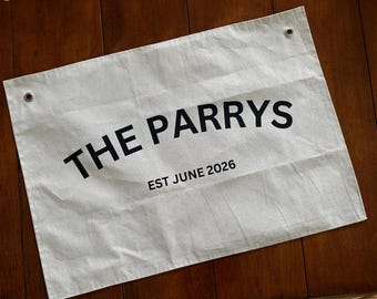 Puede incluir: Un estandarte rectangular de lona color blanco hueso con texto negro que dice THE PARRYS y EST JUNE 2026. El estandarte incluye dos ojales metálicos en las esquinas superiores para colgar, con textura de tela visible y ligeras arrugas.