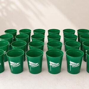 Puede incluir: Una colecci&oacute;n de veinticuatro vasos de pl&aacute;stico verde dispuestos en cuadr&iacute;cula. Cada vaso presenta un gr&aacute;fico blanco de un bander&iacute;n en un m&aacute;stil con el texto "CAMP BRIANNA" impreso en su interior. Los vasos tienen una forma cil&iacute;ndrica lisa.