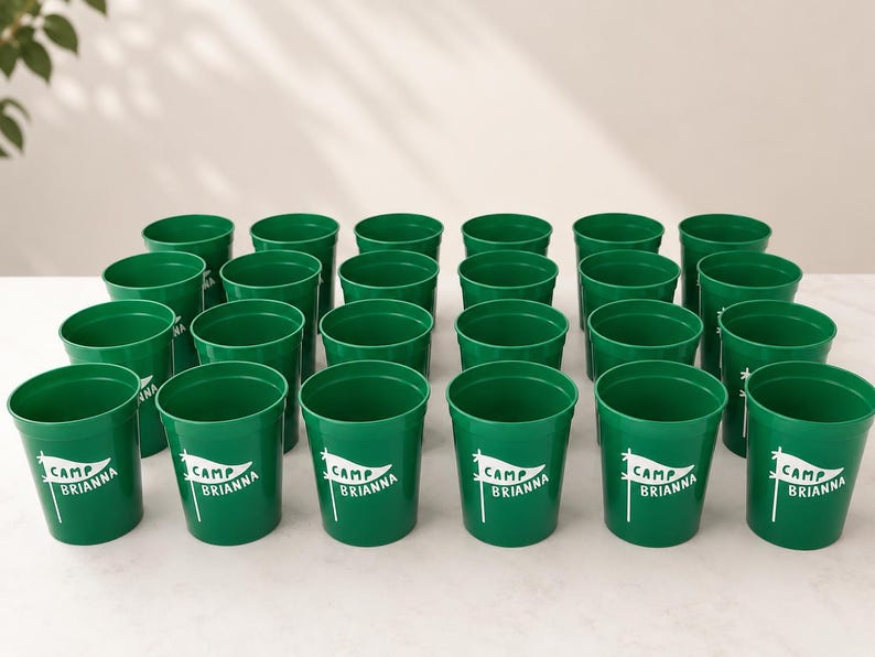 Puede incluir: Una colecci&oacute;n de veinticuatro vasos de pl&aacute;stico verde dispuestos en cuadr&iacute;cula. Cada vaso presenta un gr&aacute;fico blanco de un bander&iacute;n en un m&aacute;stil con el texto "CAMP BRIANNA" impreso en su interior. Los vasos tienen una forma cil&iacute;ndrica lisa.