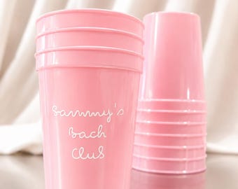 Puede incluir: Una pila de vasos de plástico de color rosa claro con el texto blanco Sammy's Bach Club impreso en letra cursiva en la parte frontal. Los vasos tienen un acabado liso y un diseño cónico con crestas horizontales cerca del borde superior.