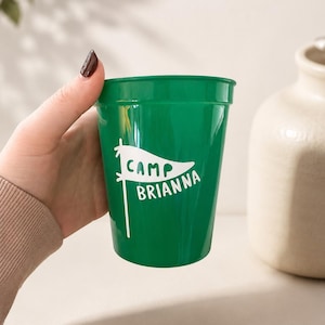 May include: A green plastic stadium cup featuring a white graphic of a pennant flag with the text "CAMP BRIANNA" printed in white lettering. The cup has a smooth, tapered cylindrical shape with a slightly flared rim at the top edge.
