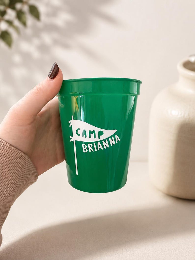 Puede incluir: Un vaso de pl&aacute;stico verde con un gr&aacute;fico blanco de un bander&iacute;n y el texto "CAMP BRIANNA" impreso en letras blancas. El vaso tiene una forma cil&iacute;ndrica lisa y c&oacute;nica con un borde ligeramente ensanchado en la parte superior.