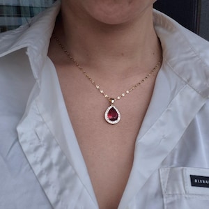May include: A gold-colored necklace with a teardrop-shaped pendant. The pendant features a large, deep red gemstone surrounded by small, clear stones. The necklace is worn over a white collared shirt.