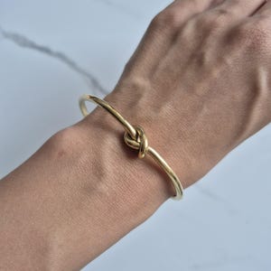 May include: A gold-coloured bracelet featuring a knot design. The bracelet is a simple, elegant piece of jewellery. The knot is the focal point, and the bracelet appears to be made of a smooth, shiny metal.