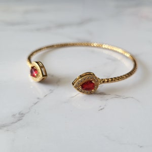 May include: A gold-colored open cuff bracelet with a textured band. Each end features a teardrop-shaped, red gemstone surrounded by small, clear stones. The bracelet is displayed on a white marble surface.