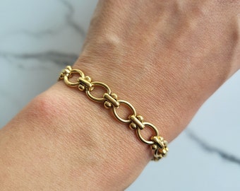 May include: A gold-colored bracelet featuring a chain-link design with oval links and decorative accents. The bracelet is made of a shiny, metallic material and is displayed on a person's wrist. The jewelry piece is a classic accessory.