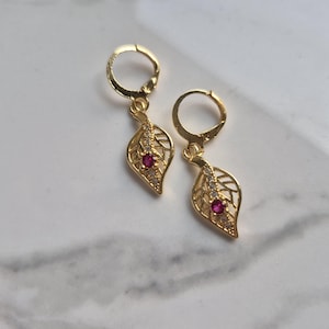 May include: A pair of gold-colored leaf-shaped earrings with a red gemstone center and small clear stones. The earrings have a hinged hoop closure and are displayed on a white and gray marble surface.