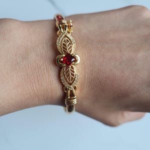 May include: Gold-toned bracelet featuring a central red gemstone and leaf-shaped filigree designs. The bracelet has a hinged clasp and is adorned with small, clear crystals. The design is symmetrical and elegant, suitable for special occasions.