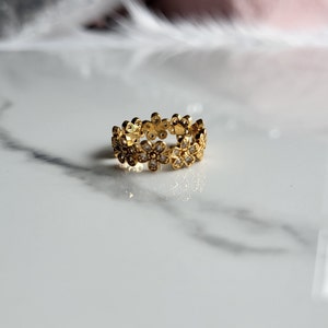 May include: A gold ring with a floral pattern. Each flower is adorned with a small, clear gemstone. The ring sits on a white and grey marble surface, which highlights the ring's warm gold colour.