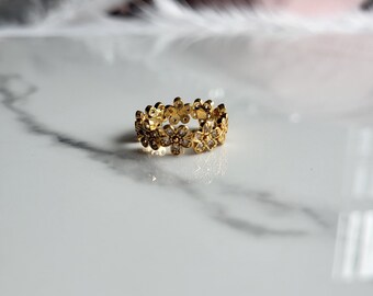 May include: A gold-colored ring featuring a floral design. Each flower has a small, clear gemstone in the center. The ring is set against a white and gray marble surface, creating a contrast with the ring's warm tones.
