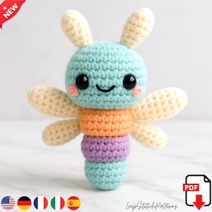 Peut inclure: Un jouet libellule au crochet fait à la main, aux couleurs pastel. La libellule présente une tête bleu clair, des segments de corps orange et violet, et des ailes de couleur crème. Le jouet a de grands yeux noirs et des joues roses. Le texte "NEW" et "LoopStitchPatterns" est inclus.