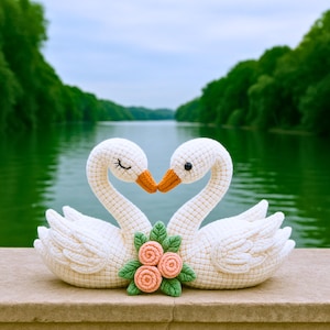 Handmade amigurumi swans in ivory yarn with layered wings, floral accents, and delicate facial embroidery, set in a light-filled bedroom.