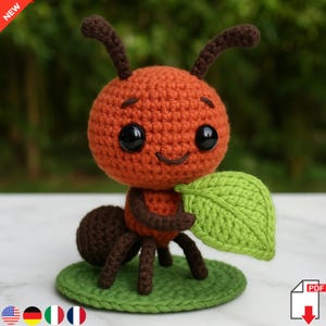 May include: A handmade crochet ant toy. The ant has an orange body, brown legs and antennae, and is holding a green leaf. It has a smiling face and is on a green base. The word "NEW" is in the top left corner.