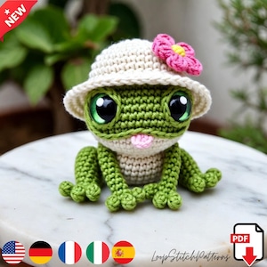 Könnte beinhalten: Eine handgefertigte Häkelfrosch mit großen, ausdrucksstarken Augen und einer kleinen rosa Zunge. Der Frosch trägt einen cremefarbenen Hut, der mit einer rosa Blume verziert ist. Der Frosch ist grün und cremefarben, und der Hut hat einen Rand. Der Frosch sitzt auf einer weißen Oberfläche.
