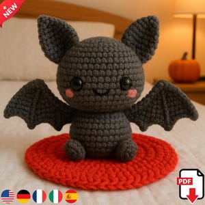 Könnte beinhalten: Eine handgefertigte Fledermaus-Häkelpuppe in Dunkelgrau mit rosa Wangen und schwarzen Augen, die auf einem roten gehäkelten Kreis sitzt. Die Fledermaus hat ausgebreitete Flügel und große Ohren. Ein "NEU"-Aufkleber befindet sich oben links.