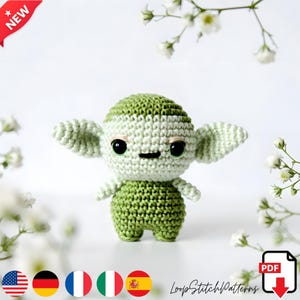 May include: A crocheted green and white doll with large ears, black eyes, and small arms and legs. The doll is surrounded by white flowers and has a "NEW" banner in the top left corner. The doll is a handmade item.
