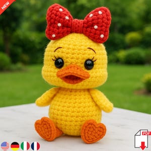 以下が含まれることがあります： 白い水玉模様の赤いリボンが付いた、手作りの黄色の編み物のアヒル。アヒルはオレンジ色のくちばしと足、大きな黒い目をしています。アヒルは緑の背景の白い表面に座っています。