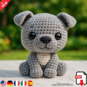 Puede incluir: Un perro de ganchillo gris hecho a mano con grandes ojos negros y una nariz negra. El perro amigurumi está sentado con las patas hacia adelante. La imagen incluye una pancarta "NUEVO" y un icono de descarga PDF.