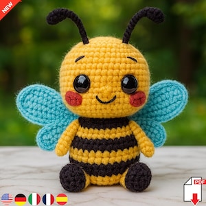 Puede incluir: Una abeja de ganchillo hecha a mano con cuerpo a rayas amarillas y negras, alas azules y antenas negras. La abeja tiene grandes ojos negros y mejillas rojas. La palabra "NEW" está en la esquina superior izquierda.