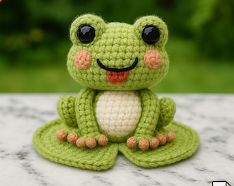 Puede incluir: Una rana de ganchillo hecha a mano en tonos verdes, con vientre color crema y mejillas color melocotón. La rana tiene grandes ojos negros y una lengua roja que sobresale. La rana está sentada en una hoja de nenúfar verde.