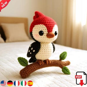 Könnte beinhalten: Ein gehäkeltes Vogelspielzeug mit rotem Kopf, schwarz-weißem Körper und braunem Schnabel. Der Vogel sitzt auf einem braunen gehäkelten Ast mit grünen Blättern. Das Design ist skurril und charmant.