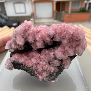 May include: A close-up of a rhodochrosite specimen. The crystal formation features clusters of translucent, pink crystals atop a dark gray matrix. The crystals have a faceted appearance, catching the light and displaying a range of pink hues.