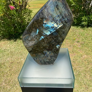 May include: A large, dark blue and green labradorite gemstone sculpture on a white pedestal with a black base. The gemstone has iridescent blue and green hues.