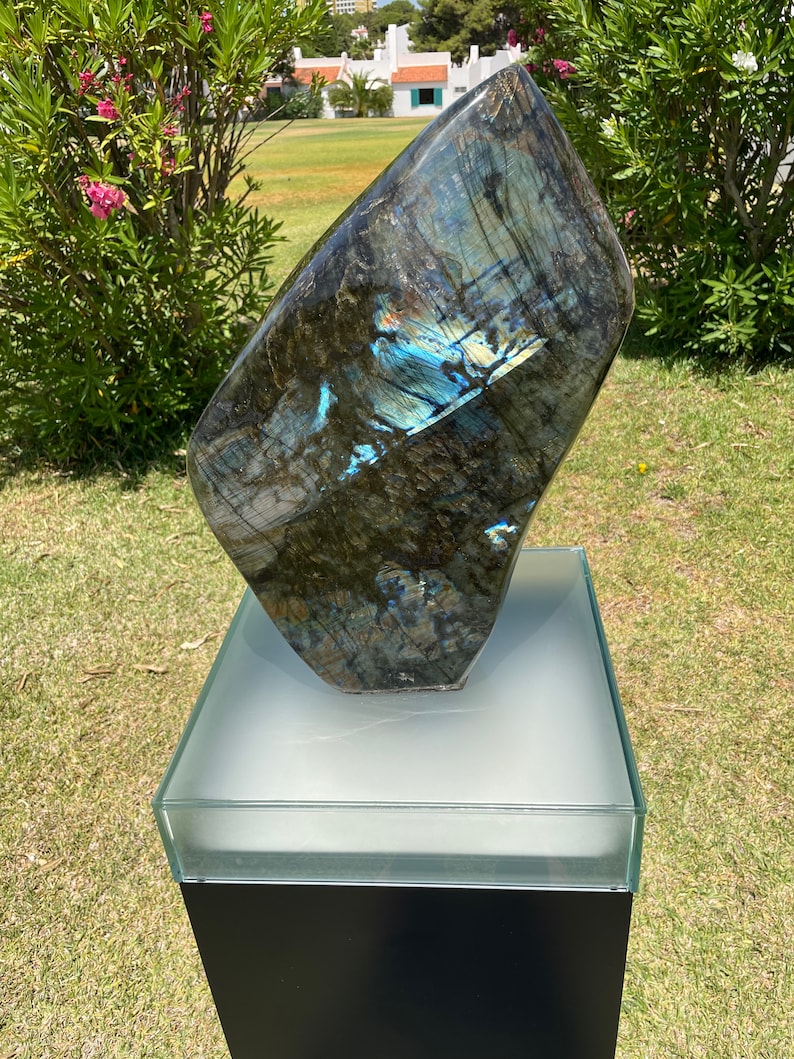 May include: A large, dark blue and green labradorite gemstone sculpture on a white pedestal with a black base. The gemstone has iridescent blue and green hues.