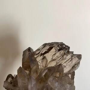 May include: A cluster of smoky quartz crystals, showcasing a range of translucent brown hues. The crystals are tightly packed, with sharp, pointed terminations. The overall shape is irregular, with a rough, textured surface. The quartz sits on a pink surface.