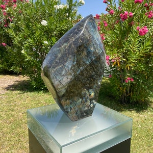 May include: A large, dark blue and green labradorite gemstone sculpture on a black pedestal with a glass top. The sculpture is shaped like a diamond and has a rough, natural texture.