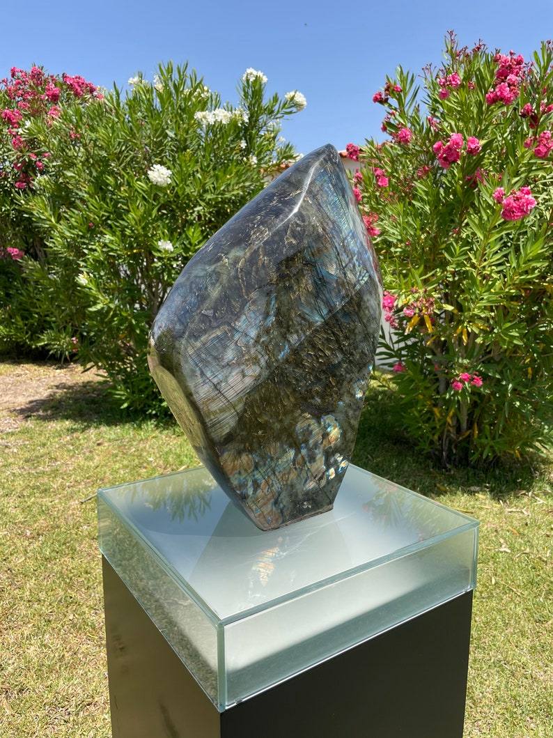 May include: A large, dark blue and green labradorite gemstone sculpture on a black pedestal with a glass top. The sculpture is shaped like a diamond and has a rough, natural texture.