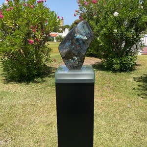 May include: A large, iridescent, blue-green labradorite gemstone sculpture on a black pedestal with a glass top. The sculpture is displayed outdoors on a grassy lawn.