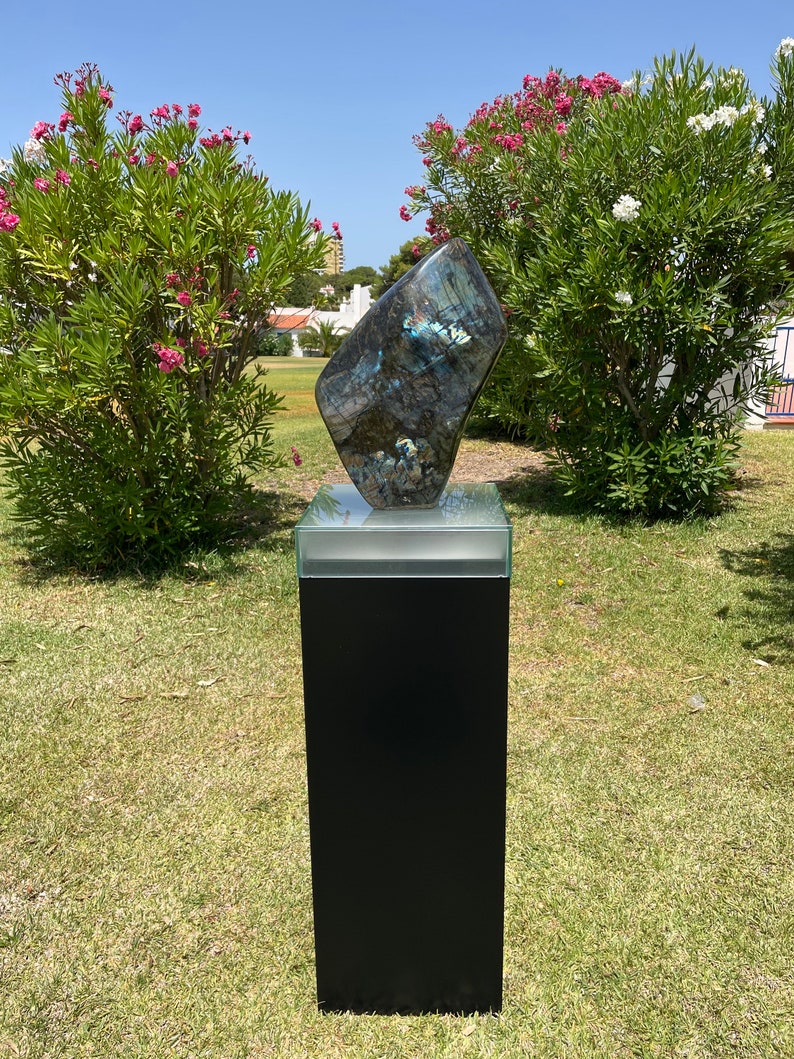 May include: A large, iridescent, blue-green labradorite gemstone sculpture on a black pedestal with a glass top. The sculpture is displayed outdoors on a grassy lawn.