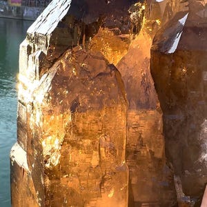 May include: A large, translucent, smoky quartz crystal cluster. The crystal has a rich, amber color with sharp, geometric facets. The background includes a body of water and graffiti-covered structures.