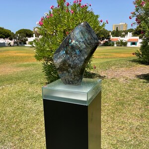 May include: A large, dark blue and green, iridescent, natural stone sculpture on a black pedestal with a glass top. The sculpture is in a grassy area with a few trees and buildings in the background.