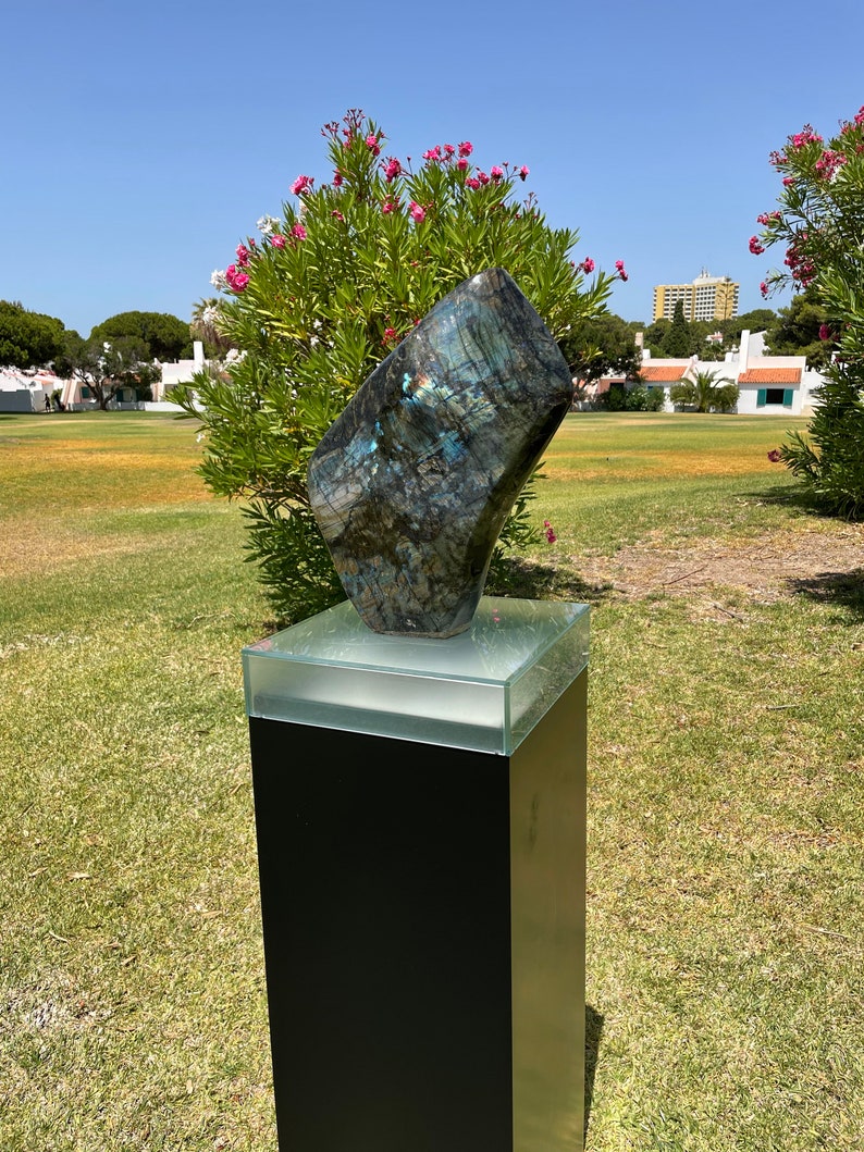 May include: A large, dark blue and green, iridescent, natural stone sculpture on a black pedestal with a glass top. The sculpture is in a grassy area with a few trees and buildings in the background.