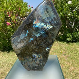 May include: A large, dark blue labradorite gemstone with iridescent blue and green flashes. The gemstone is displayed on a clear glass pedestal.