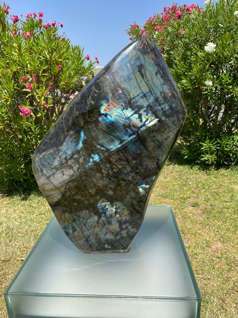 May include: A large, dark blue labradorite gemstone with iridescent blue and green flashes. The gemstone is displayed on a clear glass pedestal.