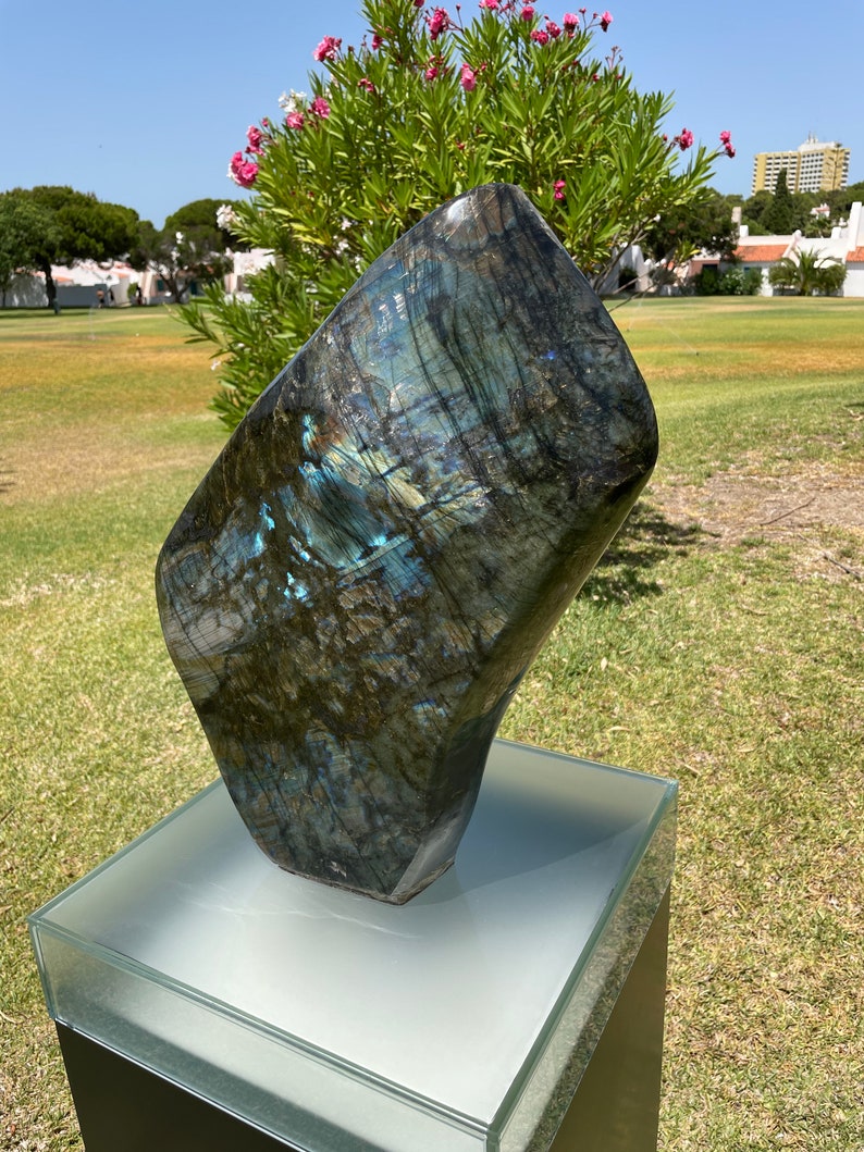 May include: A large, dark blue and green labradorite gemstone sculpture on a white pedestal. The sculpture is smooth and polished, with a unique iridescent sheen.