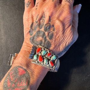 Op de afbeelding: Een zilveren manchetarmband met turquoise en rode stenen accenten. De armband heeft een gedetailleerd, sierlijk ontwerp. Een zilveren ring met een turkoois steen is ook zichtbaar.