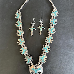 May include: A silver necklace and matching earrings set with turquoise inlay. The necklace features a series of decorative silver elements with turquoise accents, connected by silver beads. The earrings have a similar design, with a turquoise inlay.