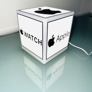 Op de afbeelding: Een witte, kubusvormige Apple Watch-display met zwarte tekst en logo's. De bovenkant heeft een zwart Apple-logo. De zijkanten tonen de woorden "WATCH" en "Apple" met het Apple-logo. Het display is verlicht en reflecteert op het oppervlak.