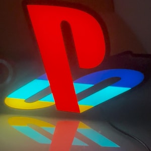 May include: A brightly lit red and blue PlayStation logo, with a yellow and blue accent, reflected on a shiny surface. The iconic symbol is illuminated, creating a vibrant display. The logo is a recognizable symbol of gaming.