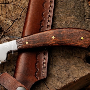 May include: Close-up of a handcrafted knife with a dark wood handle and a patterned steel blade. The knife is paired with a brown leather sheath. The handle is secured with brass rivets. The knife is resting on a wooden surface.