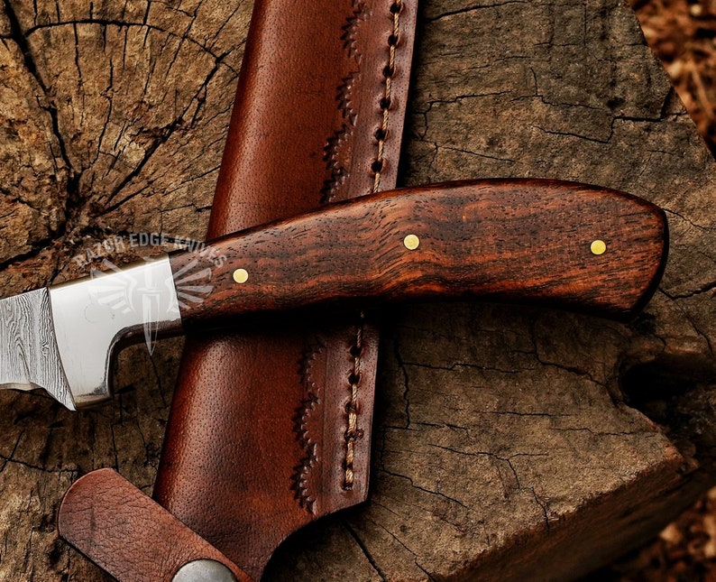 May include: Close-up of a handcrafted knife with a dark wood handle and a patterned steel blade. The knife is paired with a brown leather sheath. The handle is secured with brass rivets. The knife is resting on a wooden surface.