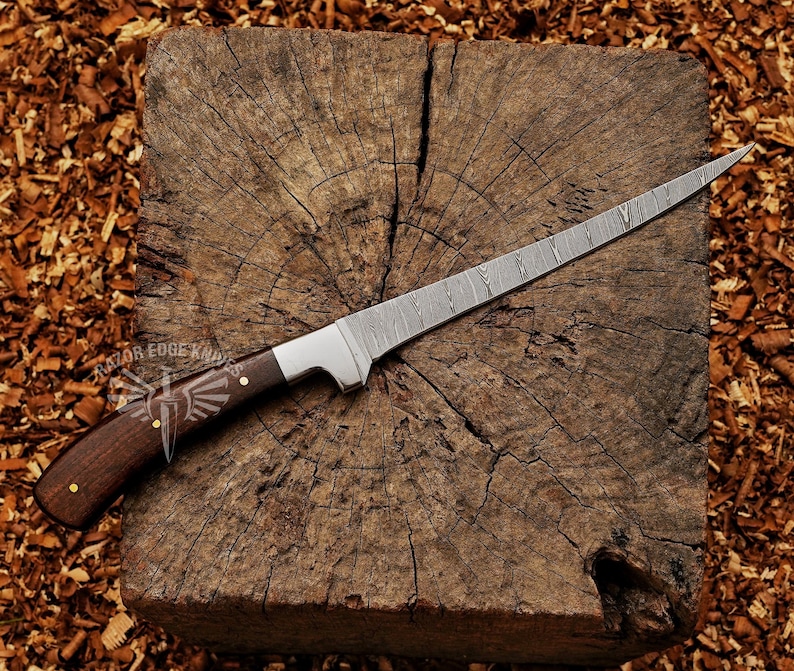 May include: A long, Damascus steel knife with a dark wood handle and a silver bolster rests on a wooden block. The handle is secured with brass rivets. The knife is designed for cutting and slicing.