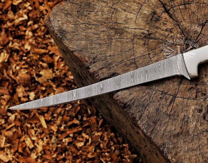 May include: A Damascus steel knife with a long, slender blade. The blade has a unique pattern and is set against a rustic wooden background. The knife's handle is white, and the text "RAZOR EDGE KNIVES" is visible.