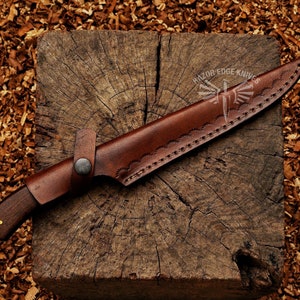 May include: A handcrafted hunting knife with a dark wooden handle and a brown leather sheath. The sheath features decorative stitching and a strap with a button closure. The knife is resting on a wooden block.
